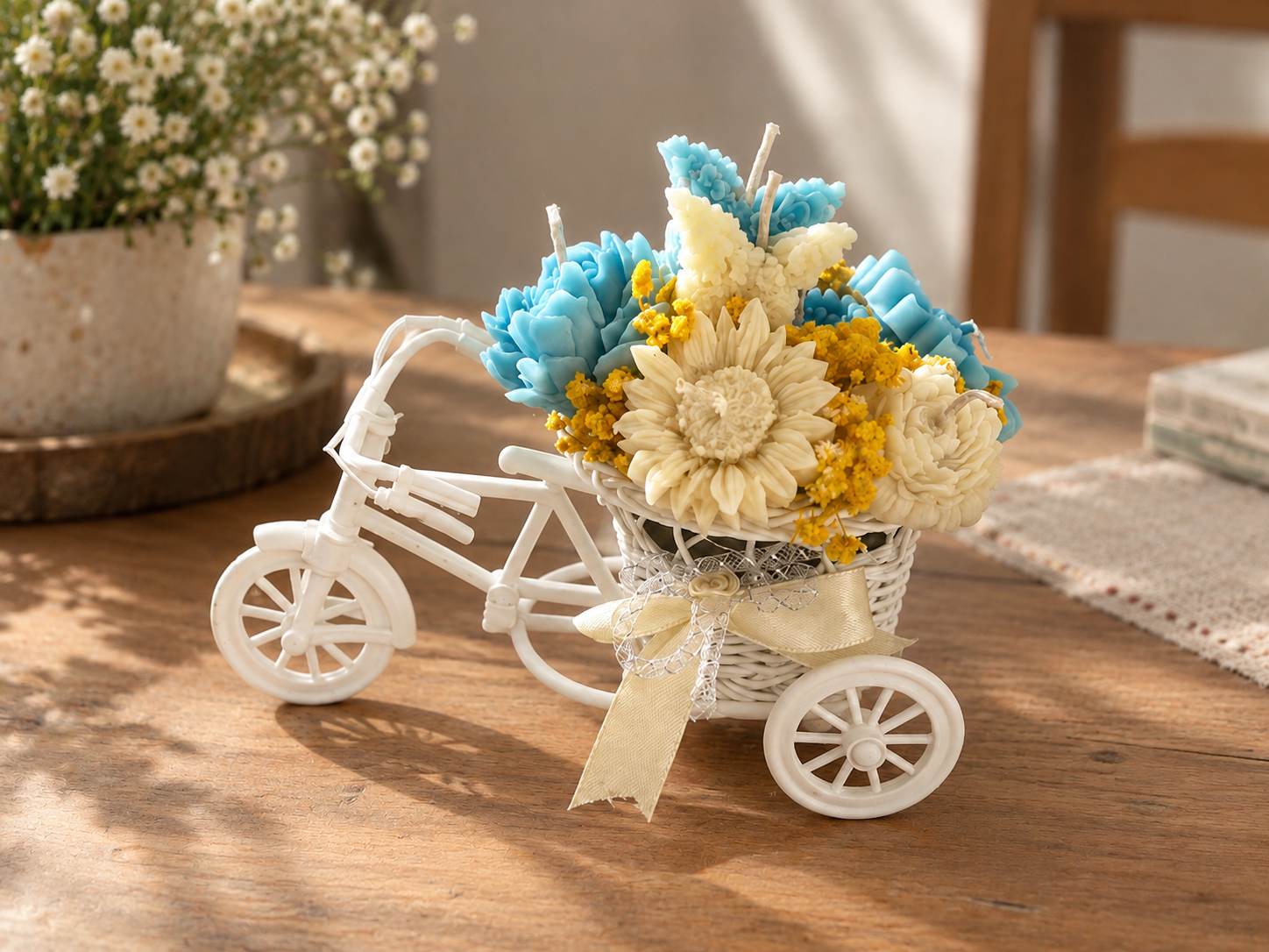 White miniature bicycle basket filled with blue and cream wax flower candles, yellow dried flowers and an ivory bow