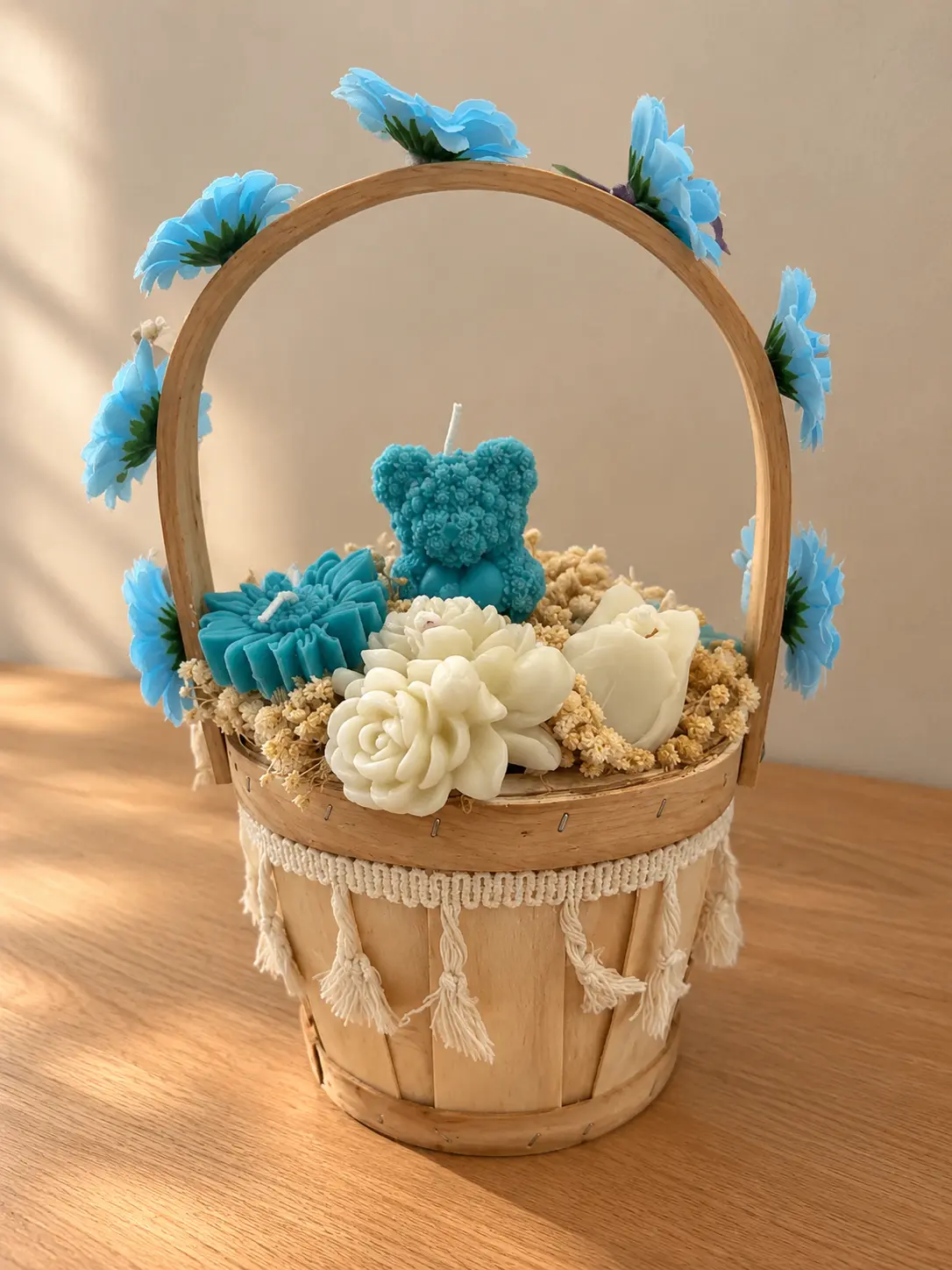 Tasselled wooden basket of teal bear and flower candles with white roses, its handle lined with blue flowers